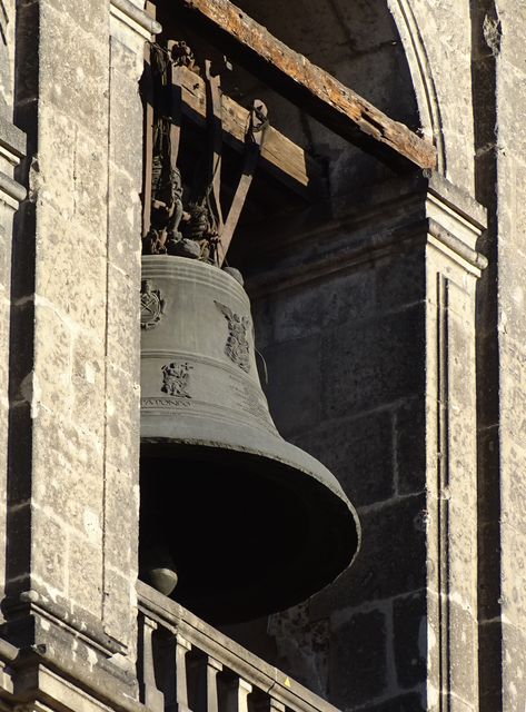 Historic bell suspended in stone bell tower with intricate masonry details. Tower partially bathed in warm sunset light emphasizing the aged texture. Suitable for use in historical articles, travel guides, cultural sites websites, and educational materials on architecture or heritage conservation.