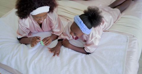 Teen sister painting younger sister's nails in pink robes on cozy bedroom bed