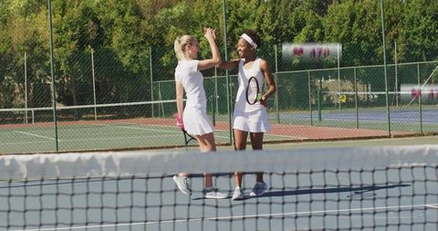 High-fiving tennis partners wearing white dresses on outdoor court celebrating victory