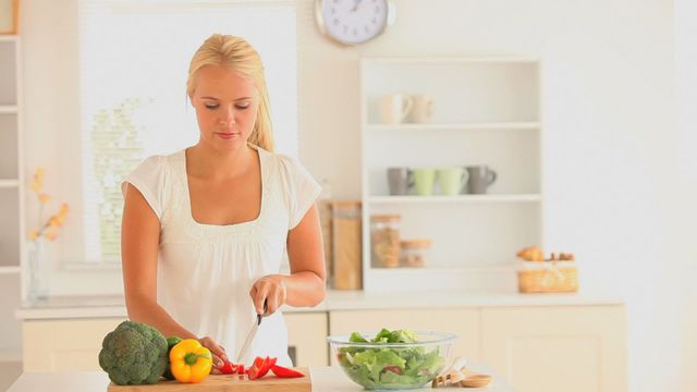 Blonde woman slicing vegetables in a bright, modern white kitchen. Showcases healthy eating and home cooking. Can be used in articles focusing on healthy lifestyle, meal prep, cooking tutorials, or kitchen design.