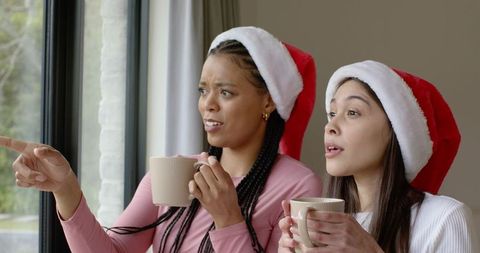 Diverse friends wearing santa hats holding mugs and pointing by window, cozy holiday