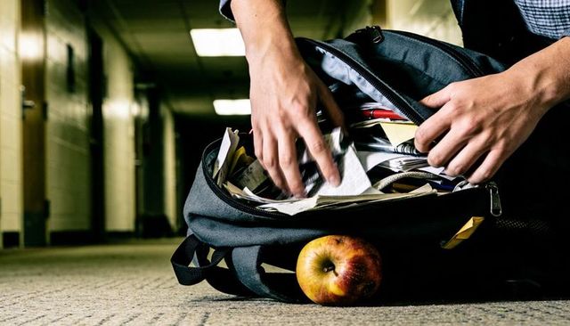 Student searching through cluttered backpack on school hallway floor under moody fluorescent lightin