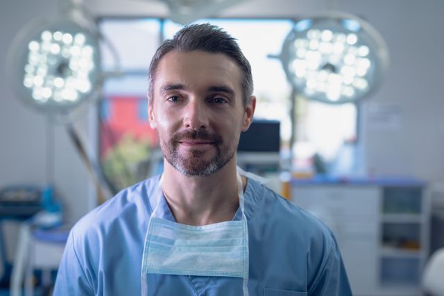 Male surgeon standing confidently in a well-lit operation room, wearing a surgical mask and scrubs. Ideal for use in medical articles, healthcare websites, hospital brochures, and educational materials about surgery and medical professions.
