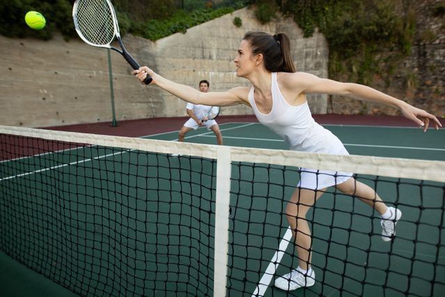 Front view of a Caucasian woman and a man wearing tennis whites playing tennis, the woman hitting the ball the man standing in the background.