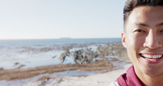Smiling young man enjoying day at rocky beach, sea in background, showcasing happiness and youth. Ideal for promoting travel, outdoor activities, summer adventures, and lifestyle content.
