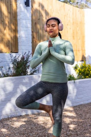 Asian woman practicing outdoor yoga tree pose wearing pink headphones and activewear