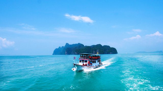 Tourist boat filled with passengers is sailing across the vibrant blue sea, leaving trails in its wake. The boat is heading towards picturesque tropical islands under a clear, sunny sky, suggesting a day filled with adventure and exploration. This can be used for travel brochures, websites promoting tropical vacations, maritime adventures, or island tourism.