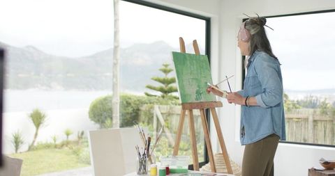 Mature woman painting on easel in bright coastal studio with large windows and palette