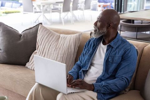 Senior african american man working on laptop relaxing on beige sofa in bright living room