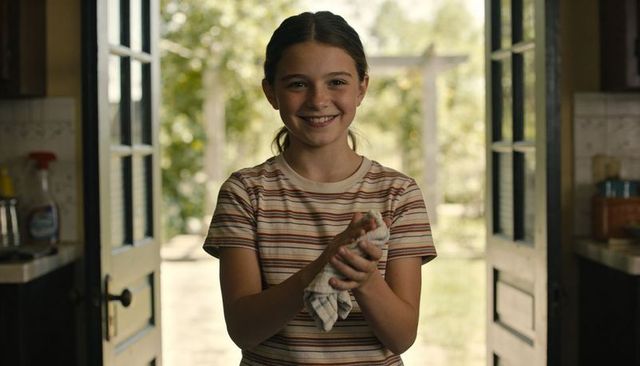 Smiling preteen girl holding dishcloth in sunlit kitchen doorway, striped tee, garden view