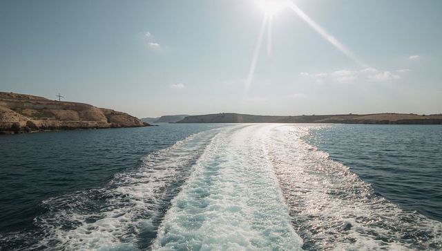 Central boat wake cutting through blue sea toward sunlit coastline