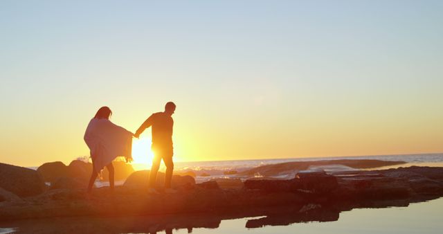 A young man and woman enjoy a romantic moment by the sea at sunset, with copy space. Their silhouettes against the vibrant sky create a peaceful and intimate atmosphere.