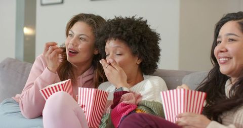 Multicultural women laughing and snacking on sofa during cozy friends movie night