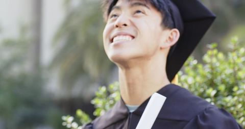 Joyful asian graduate wearing mortarboard and gown holding diploma on sunlit campus