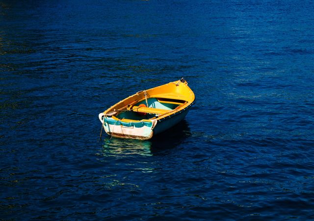 Small, brightly colored rowboat floating on calm, deep blue water. This serene and tranquil scene can be used for relaxation themes, travel brochures, peaceful nature presentations, or as a background image depicting solitude.