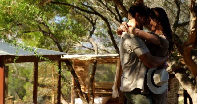 A young Caucasian couple shares a romantic embrace outdoors, surrounded by trees and a wooden structure, with copy space. Their affectionate moment captures a sense of intimacy and connection with nature.