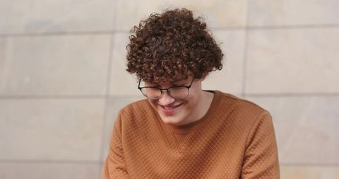 Smiling young man looking down wearing round glasses and rust pullover on beige tile wall