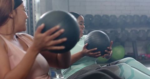 Diverse women rotating torsos holding medicine balls during partner core workout in gym