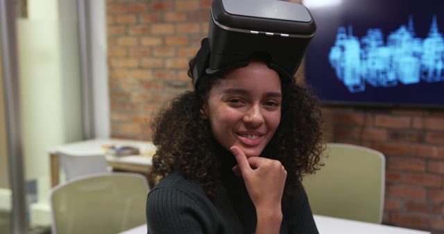 Smiling woman wearing a virtual reality headset in a modern office. Shows technology adaptation in casual work environments. Ideal for articles on VR tech, modern workplaces, startups, and innovation. Useful for website headers, tech forums, and promotional materials highlighting futuristic office settings.