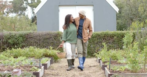 Diverse couple walking through backyard raised beds carrying wicker basket and smiling