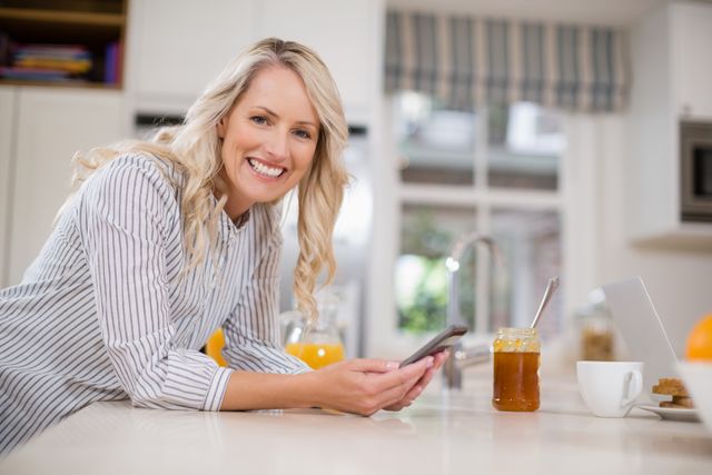 Woman leaning on kitchen counter, using mobile phone and smiling. Ideal for lifestyle blogs, technology, domestic life articles, home decor inspiration, and modern living promotions.