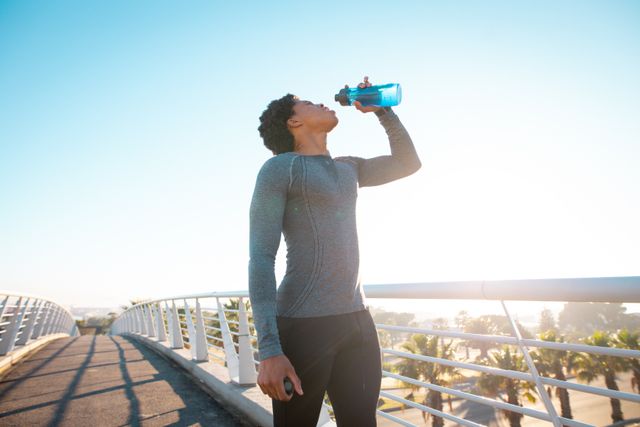 Fit man drinking water while exercising outdoors on a bridge in the city. Ideal for promoting healthy lifestyles, fitness routines, hydration importance, and urban exercise. Suitable for use in fitness blogs, health magazines, and sportswear advertisements.