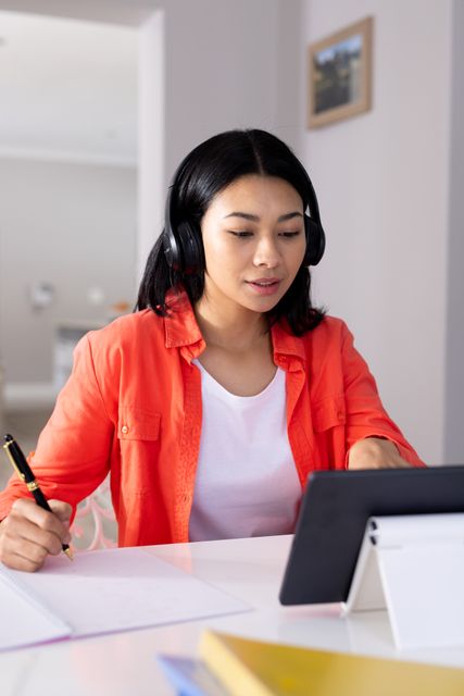 Biracial woman wearing headphones, writing notes while participating in a video call on a tablet at home. Ideal for illustrating remote work, modern communication, and home office setups. Useful for articles on work-life balance, technology in daily life, and virtual meetings.