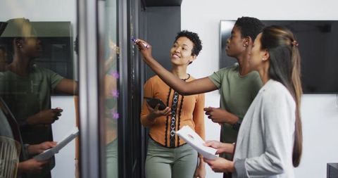 Diverse businesswomen collaborating at glassboard in modern office