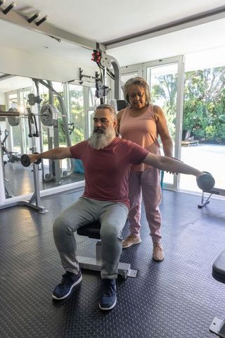 Senior couple working out in home gym, man lifting dumbbells while partner spotting
