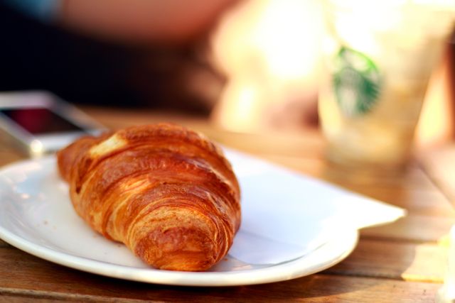 Croissant on white plate next to iced coffee cup and smartphone on wooden table. Perfect for illustrating breakfast, morning routines, or cafe scenes. Ideal for use in food blogs, restaurant menus, and lifestyle advertisements.