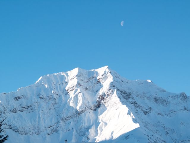 Captivating view of snow-covered mountain peaks with a bright blue sky and the moon overhead. Perfect for use in travel brochures, nature magazines, winter sports advertisements, and as a serene landscape background. The scene captures the calmness and beauty of nature, ideal for inspiring wanderlust and outdoor adventure.