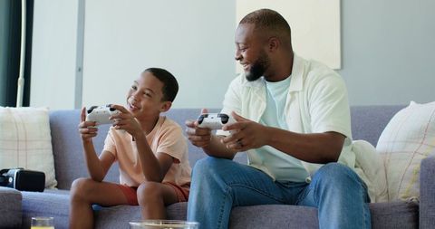 African american father and son playing video games on sofa sharing controllers