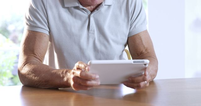 An elderly man sits at a wooden table, using a tablet for browsing the internet or entertainment. The setting appears to be the comfort of his home. This image is suitable for websites, articles, and advertisements focusing on senior lifestyle, digital literacy, technology for older adults, and comfortable living for the elderly.