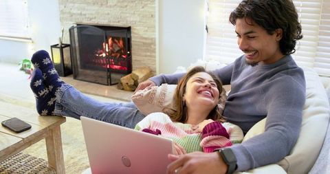 Couple lounging on sectional sofa with laptop by fireplace, sharing warm cozy moment