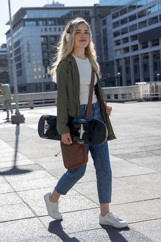 Young woman walking with skateboard and headphones in urban plaza, casual street style