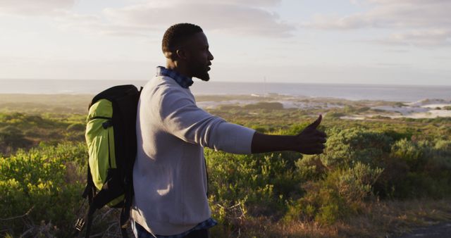 African american man with backpack trying to hitch a lift while standing on the road. road trip travel and adventure concept