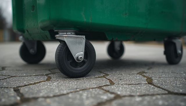 Heavy-duty rubber caster wheel rolling under green commercial waste container on paver stones