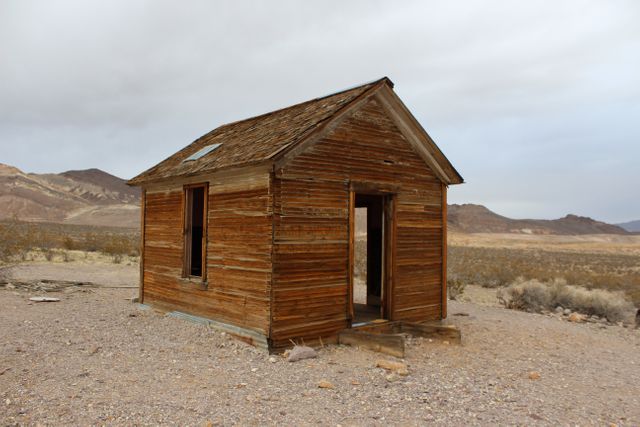 This image shows a weathered and abandoned wooden cabin in the midst of a vast and desolate desert. The landscape is dry, barren, surrounded by mountains in the background, highlighting isolation and solitude. This image could be used for themes of abandonment, history, rustic architecture, and the beauty of nature reclaiming human structures. It is suitable for illustrating articles or stories about exploration, survival in harsh environments, and the passing of time.