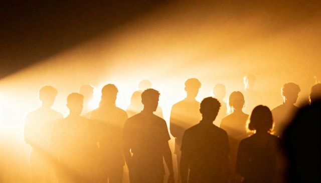 Standing silhouetted crowd bathed in golden backlight with dramatic beams and theater haze