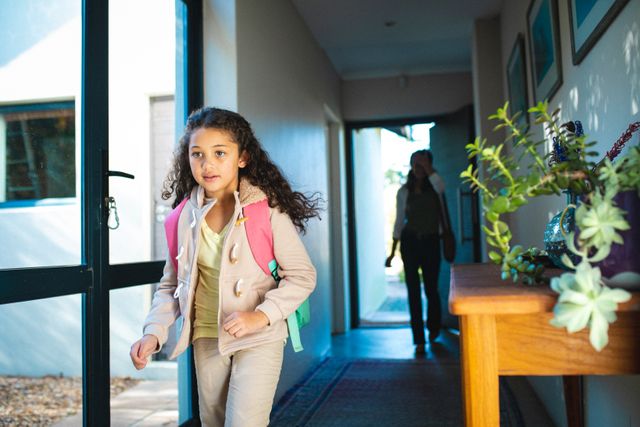 Young biracial girl with curly hair wearing a backpack, walking through hallway towards door, ready for school. Parent visible in background. Ideal for illustrating themes of education, morning routines, family life, and childhood. Suitable for use in educational materials, parenting blogs, and lifestyle articles.