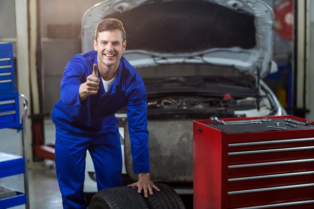Mechanic in blue overalls smiling and showing thumbs up while standing next to a car with an open hood and a tire in a repair garage. A red toolbox is visible in the foreground. Ideal for use in advertisements for automotive services, repair shops, and car maintenance tutorials.