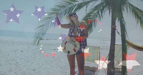 Blonde woman relaxing on tropical beach holding straw hat under palm with decorative stars