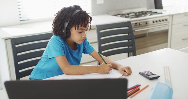 Young boy is doing homework in a modern kitchen, wearing headphones, sitting at a table with a calculator and other school supplies. Ideal for use in educational websites, online learning platforms, and articles about children's education and study habits.