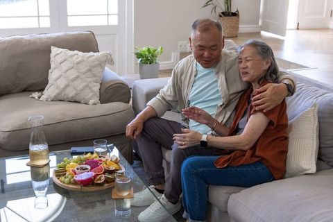 Senior asian couple sitting on sofa sharing fruit platter and smartphone in sunlit home