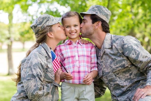Military parents in camouflage uniforms kiss their daughter who is holding an American flag in a park on a sunny day. This image can be used to depict family bonding, patriotism, support for military families, and outdoor activities.