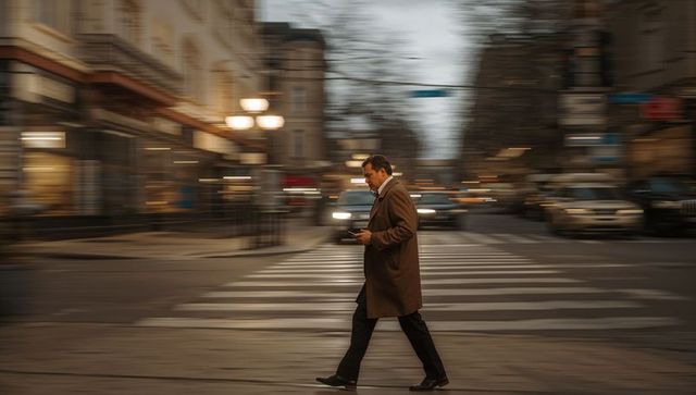 Businessman crossing city street at dusk while checking smartphone with motion blur