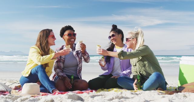 Four women of different ethnicities are sitting on a sandy beach, smiling, and enjoying drinks together under the sunshine. They appear relaxed and happy, making this perfect for promoting themes of friendship, leisure, or summer vacations. This can be used in travel advertisements, social media posts about summer plans, lifestyle blog articles, or advertisements for beachwear.