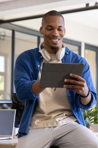 African american man smiling using tablet in modern office wearing headphones and denim