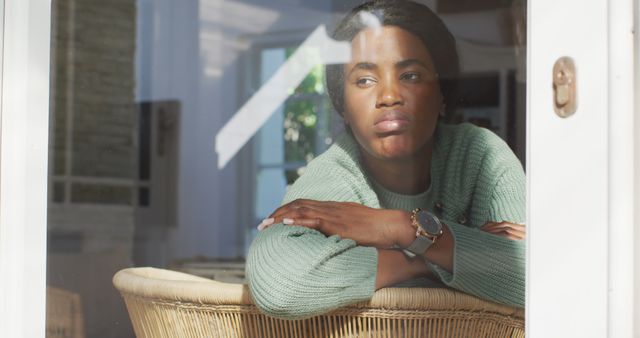 A woman seated at an indoor space looks through the window pensively. She is wearing a green sweater and a wristwatch, highlighting a blend of casual fashion and reflective mood. Useful for illustrating concepts of contemplation, relaxation, thoughtfulness, and everyday life moments.