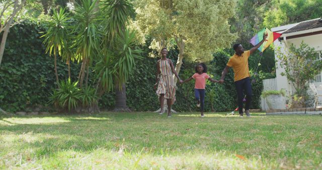 Photo of a joyful African American family spending quality time together in a lush green yard, flying a colorful kite. Mother, father, and daughter holding hands, smiling and running. Ideal for use in advertisements promoting family activities, outdoor leisure, summer fun, and togetherness themes.
